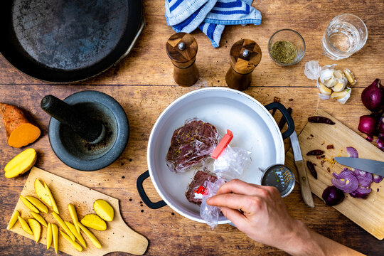 Preparation of raw beef tenderloin steak for sous vide bath in pot with thermometer on wooden table with steel pan,  raw potato wedges rosemary, chili, onion and garlic, photographed from above.