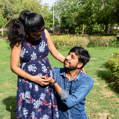 Indian couple posing for maternity baby shoot. The couple is posing in a lawn with green grass and the woman is falunting her baby bump in Lodhi Garden in New Delhi, India