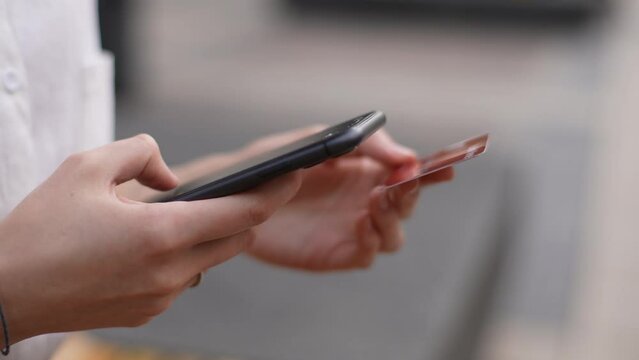 Close-up hands of unrecognizable young woman holding bank card, entering cvv code or payment information in mobile internet shopping application, standing on city street. Shooting in slow motion.