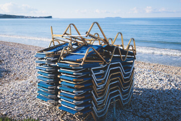 Stack of sunbeds chaise-longues piled up on the beach in the end of touristic season in Greece, Ionian sea islands - obrazy, fototapety, plakaty