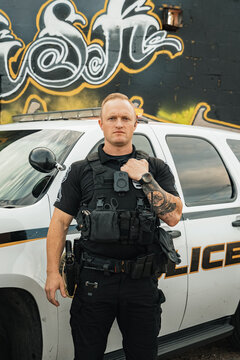 Vertical image of white male caucasian police officer posing with straight face and one hand on bulletproof vest standing in front of his cop car with graffiti wall backdrop.
