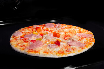 Uncooked pizza with sausage, ham, mushroom, pepper, cheese on tray in electric oven, black background. Italian cuisine, homemade bakery, fast food, cooking, semi-finished products and raw concept