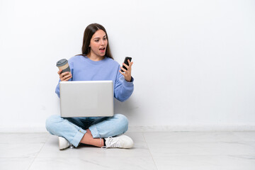 Young caucasian woman with laptop sitting on the floor isolated on white background holding coffee to take away and a mobile