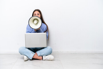 Young caucasian woman with laptop sitting on the floor isolated on white background shouting through a megaphone