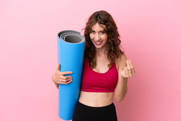 Young sport woman going to yoga classes while holding a mat isolated on pink background making money gesture