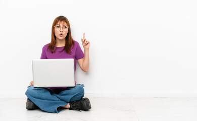 redhead girl with a laptop sitting on the floor thinking an idea pointing the finger up