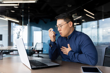 Young male Asian office worker having asthma attack at workplace, breathing hard. He sits at a...