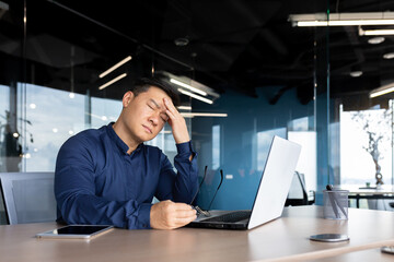 A young Asian man, an office worker, feels a strong headache. Migraine. He sits at the desk, holds his head, closed his eyes, took off his glasses, and rests.