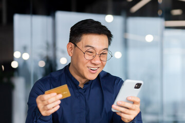 Close-up photo. A happy young Asian man in glasses holds a credit card and looks joyfully at the phone. Checks the account, makes online purchases, orders.