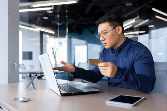 Worried young Asian man sitting in the office at the table, holding a credit card. Looking shocked at the laptop monitor, spreading his hands. Checks account, failed online purchases, bankruptcy.