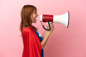 Super Hero redhead girl isolated on pink background shouting through a megaphone