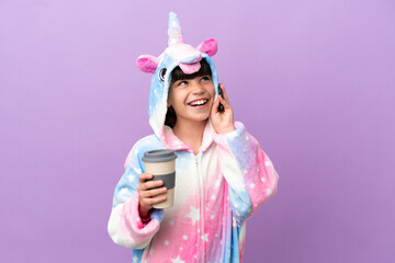 Little kid wearing a unicorn pajama isolated on purple background holding coffee to take away and a mobile