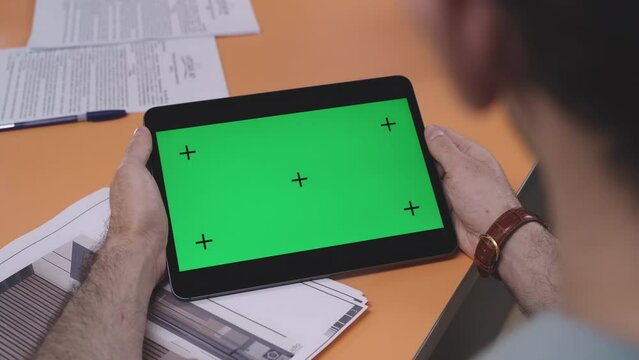 Businessman holds tablet computer with blank chromakey screen at table in office close rear view. Communication technology