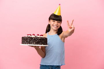 Little caucasian kid holding birthday cake isolated in pink background smiling and showing victory...