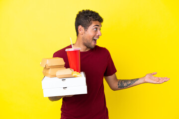 Young brazilian man holding pizzas and burgers isolated background with surprise expression while looking side