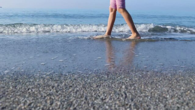 Calm morning at sea. Little girl's legs walking along seashore from right to left side of frame. Vacation, tourism concept.