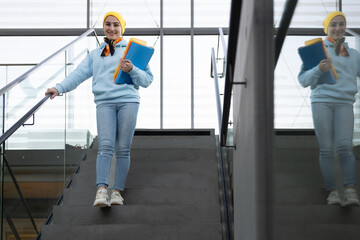 Smiling female student with multi-colored folders in her hands goes down the stairs