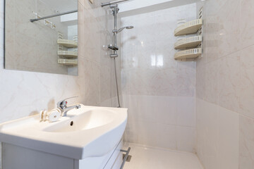 Small bathroom with a white porcelain sink on a wooden cabinet and a frameless square mirror, a plastic shelf in one corner and a walk-in shower without a screen