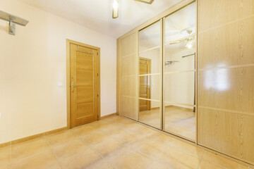 Empty room with wall-to-wall built-in wardrobe with oak sliding doors and mirrored ceiling fan, cream painted walls and light brown tiled floors