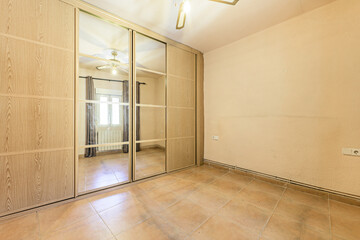 Empty room with wall-to-wall built-in wardrobe with oak sliding doors and mirrored ceiling fan, cream painted walls and light brown tiled floor