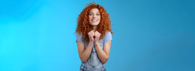 Pretty please buy me that. Amused longing excited redhead cheerful girl clench fists together stare camera desire express aspiration wanna receive buy product standing blue background thrilled - obrazy, fototapety, plakaty