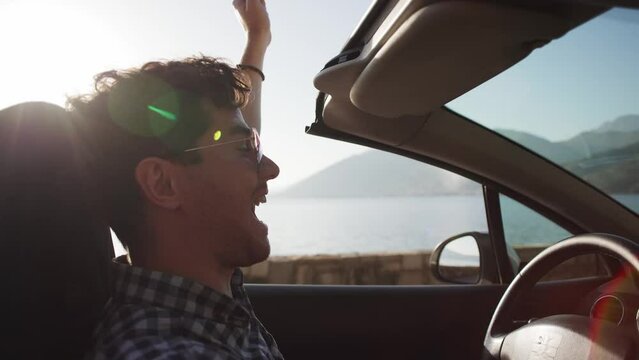 A young men in a shirt rides a convertible along the seashore in the mountains, enjoys the wind with his hand. Vacation, car purchase and car rental