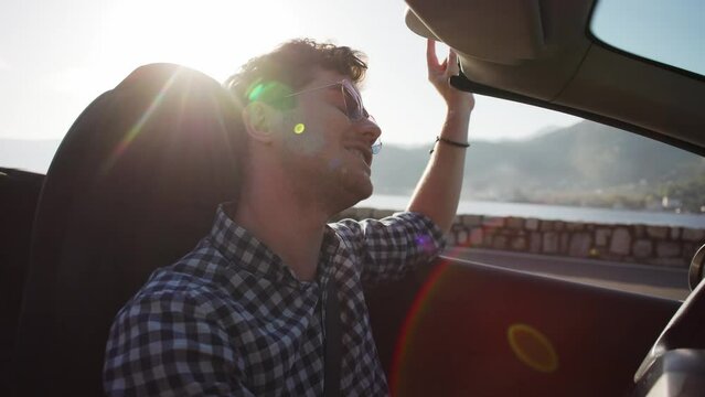 A young men in a shirt rides a convertible along the seashore in the mountains, enjoys the wind with his hand. Vacation, car purchase and car rental