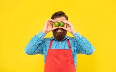 Happy guy in apron holding fresh limes on eyes yellow background, fruit seller