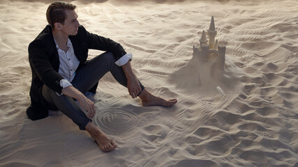 Financier and sand castles, the concept of unprofitable investments. Economic crisis and recession. A young man in a business suit is sitting on the sand next to a sand castle - obrazy, fototapety, plakaty