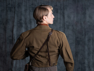 A young guy in military-style clothes, a brown flight jacket and breeches with suspenders. Posing in the studio on a gray background, back view