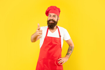 Happy man in toque and apron pointing finger ahead yellow background, cook