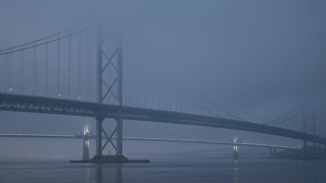 A large suspension bridge over the sea bay covered with fog or haar. The lights of cars moving across the bridge. Video with the panning camera motion. Forth Road Bridge, Scotland, United Kingdom