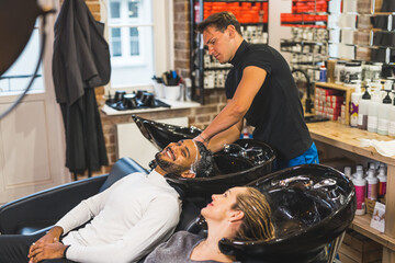 Male hairdresser washing hair of his clients at beauty salon. Indoor shot. Happy couple visiting...