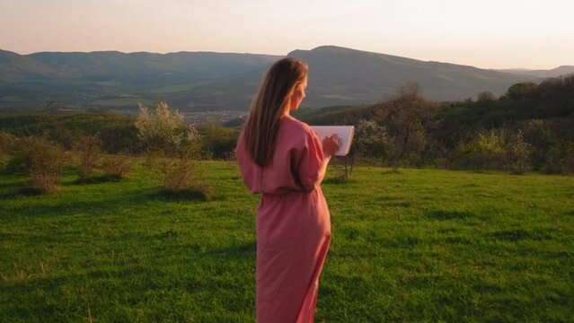 the girl stands against the background of a beautiful landscape at sunset and draws. a girl paints at sunset. the artist paints a landscape. En plein air painting. sketch of the landscape