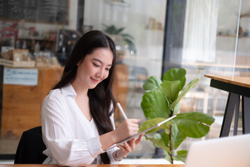 Portrait of young beautiful asian woman holding stylus pen, thinking and looking outside while...