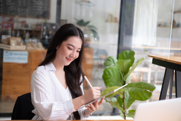 Portrait of young beautiful asian woman holding stylus pen, thinking and looking outside while...