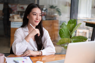 Portrait of young beautiful asian woman holding stylus pen, thinking and looking outside while...