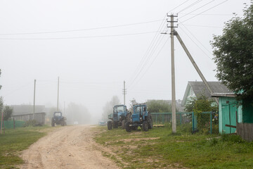 Misty morning in the countryside. View of a deserted village street. Old tractors on the side of a dirt road. Foggy rural landscape. Everyday life in the countryside. Vologda region, Russia.