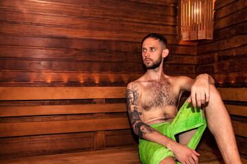 Young bearded man in green towel sitting on bench in bathhouse sauna and relaxing, looking away. Guy resting in finnish sauna at wooden wall. Wellness, self care, healthy concept. Copy text space