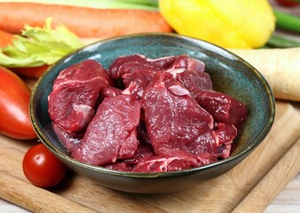 Raw deer meat for venison ragout or goulash. Bowl with pieces of deer meat on cutting board, fresh vegetable for cooking around.