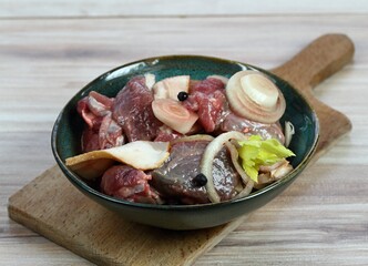 Fresh deer meat  marinated in red wine with bakon, onion, garlic and juniper berries. Bowl with pieces of deer meat on cutting board, wooden table.