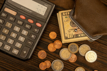 Dollar banknotes, euro coins, a vintage brown leather wallet and a calculator on a wooden surface.