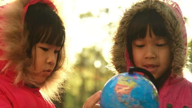 Two cute Asian girls learning a model of the world on nature background and warm sunlight in the park. Children learn through educational play activities. Earth day. World environment day.