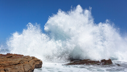 Wave Rocks Exploding Crashing Breaking White Water Spray Against Blue Sky Along Rocky Beach Coastline.