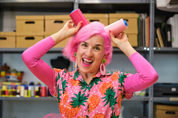 Dressmaker with pink hair and colorfull clothes making funny faces with sewing thread reels in a sewing workshop.