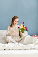 Happy woman sitting on the bed wearing pajamas holding tulip flowers bouquet chatting using laptop showing thumb us