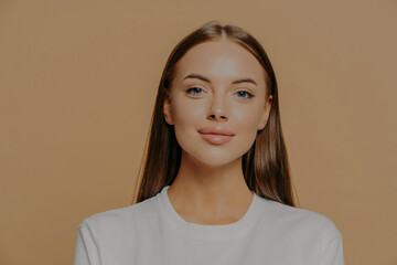 Portrait of pretty European woman with long hair looks confidently ar camera has healthy glowing skin makeup wears casual jumper isolated over brown studio wall. Beauty and wellness concept.