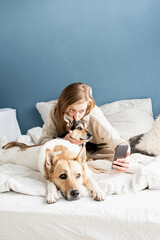 Beautiful young adult cheerful woman taking selfie on smartphone with her dog in morning in bed