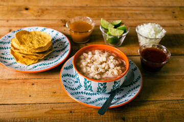 Pozole served in a deep dish, with tostadas, salsa and vegetables on a wooden table. Typical Mexican food.
