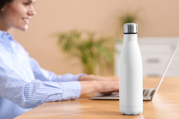 Young woman with laptop at table indoors, focus on thermo bottle